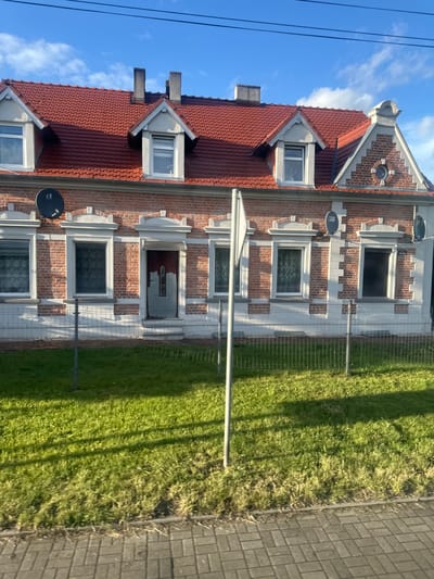The image captures a two-story brick building with a red tile roof, featuring multiple dormer windows. The building has a classic facade with decorative brickwork and stone accents around the windows. A dark-colored front door is visible, partially obscured by a silver awning. The property is enclosed by a metal fence, and a patch of green grass with some dry vegetation runs along the front. In the foreground, a paved sidewalk or road is visible. The sky above is blue with scattered white clouds. The scene appears to be an exterior view of a residential or commercial property, likely in Krosno Odrzańskie, Poland, given the context. There are no people visible in the image. Satellite dishes are mounted on the exterior of the building. A street number "183 443" is visible near one of the windows.