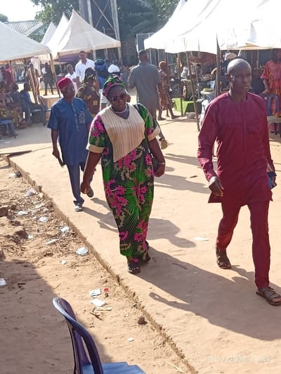This image depicts an outdoor gathering in Isinyi-Nando, Nigeria, occurring on 2025-12-19 at 15:41 local time. The scene is illuminated by direct sunlight, casting distinct shadows.
In the foreground, a group of three individuals walks along a dirt path. A woman, positioned centrally, wears a green, pink, and black patterned dress with a matching headwrap, sunglasses, and a white necklace, carrying a dark handbag. She is walking towards the viewer. To her left and slightly behind, a man in a blue long-sleeved tunic with embroidery and a red cap walks in the same direction, wearing dark sandals. To the woman's right, a man in a maroon long-sleeved tunic and matching trousers walks towards the viewer, wearing sandals.
The ground is composed of compacted dirt, with a slightly raised curb or edge running parallel to the path. Along this edge, debris including white paper and plastic fragments is scattered. A blue plastic monobloc chair is partially visible in the lower-left corner of the frame.
In the mid-ground and background, multiple white canopy tents are erected, sheltering numerous individuals. People are seen standing and seated under these tents, dressed in various garments. A tall, dark, industrial-looking structure, possibly a water tank or tower, is visible behind the tents on the left side. Additional structures and sparse vegetation are present further in the background.