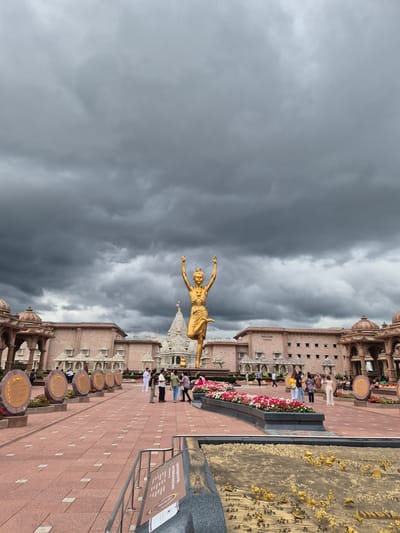 The image depicts the exterior of a large, ornate temple complex under a dark, cloudy sky in Robbinsville Township, New Jersey, United States of America. In the foreground, a golden statue of a person in a dynamic pose stands prominently on a raised platform. The statue is gold and appears to be a significant cultural or religious figure.
Surrounding the central statue is a vast, paved courtyard with intricate tile patterns. Several people are visible walking and conversing in the courtyard, dressed in casual attire. To the left and right of the courtyard are elaborate architectural structures with domes and intricate carvings, characteristic of Indian temple architecture. Some of these structures appear to be entrances or pavilions.
Behind the courtyard and the statue, a long, multi-story building with arched windows is visible. A sign on this building reads "WELCOME CENTER". Further in the background, another white, multi-tiered structure with a spire can be seen, possibly another part of the temple complex.
Along the edge of the courtyard, there are circular plaques or exhibits displayed on stands, some of which are surrounded by vibrant flower beds. In the far right foreground, a detailed miniature model of a scene, possibly a historical or mythological event, is displayed within a rectangular structure with golden figures and elements. The overall atmosphere is one of grandeur and cultural significance, contrasted by the looming, overcast sky.