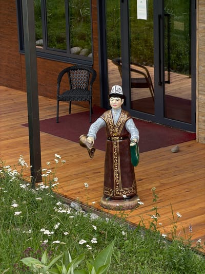 This is a vertical, eye-level medium shot of a life-sized statue of a person dressed in traditional Kyrgyz attire. The statue stands on a wooden deck in front of a building with large glass doors, possibly an entrance. The figure is a man wearing a brown, embroidered robe with gold trim, a white shirt with light blue sleeves, and a traditional white and black patterned hat. He is holding a green object, possibly a bag or slipper, in his left hand.

To the left of the statue is a black wicker chair. Beyond the doors, the reflection shows green grass and trees, suggesting an outdoor setting. A red mat is visible on the deck in front of the doors. The wooden planks of the deck are laid out horizontally in the frame. To the right of the statue, a patch of green grass with small white wildflowers grows, bordering the deck.

The lighting suggests it is daytime, with natural light casting shadows on the deck. The scene appears to be calm and perhaps welcoming, given the depiction of a traditionally dressed figure and the inviting outdoor space. The overall impression is one of cultural representation and hospitality. No visible text is apparent in the image or its crops. The location context is Kaindy, Kyrgyzstan.