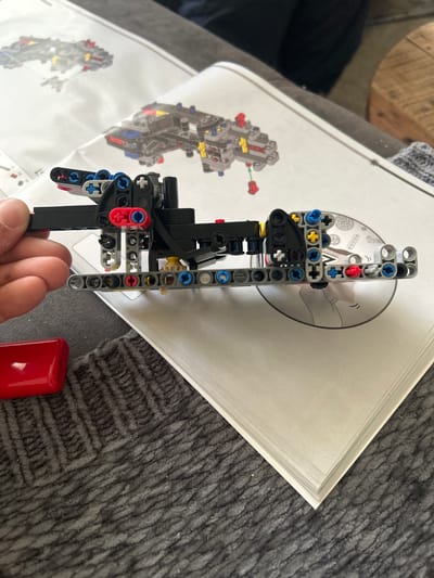 The image displays a close-up view of a person's hand holding a partially assembled LEGO Technic model. The model consists of various colored plastic bricks, gears, and connecting pins. In the background, an instruction booklet for the LEGO set is visible, showing diagrams of the model. A red rectangular object is positioned on a dark gray, textured surface, likely a rug or blanket, to the left of the LEGO model. The scene appears to be indoors, with the background suggesting a domestic environment. There are no discernible landmarks or elements that would indicate a specific location in Leiden, Netherlands.
