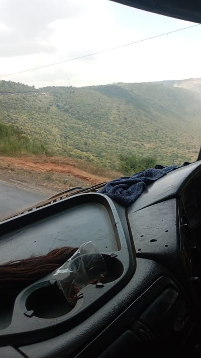 The image depicts a panoramic view from the interior of a vehicle, facing outwards towards a mountainous landscape in Lari/Kirenya ward, Kenya. In the immediate foreground, the dark, textured plastic dashboard is visible, featuring a flat area with two circular cup holders. On the dashboard, a length of dark reddish-brown synthetic hair is present, along with a clear plastic bag containing small, reddish-brown spherical objects, positioned within or adjacent to a cup holder. A dark blue, folded towel rests on the passenger side of the dashboard near the base of the windshield. The lower portions of black windshield wipers are visible at the bottom of the windshield.
Beyond the vehicle, a paved, two-lane road, dark grey in color, curves along the base of a reddish-brown dirt embankment on the left side of the frame. The midground and background are dominated by an expansive series of rolling hills and mountains, densely covered in light green, scrub-like vegetation and trees. Patches of reddish-brown earth are visible on the slopes where vegetation is sparser. In the distant background, a tall, slender mast or tower is visible atop a ridge. To the far right, a faint, light-colored plume of dust or smoke rises above the distant hills. The sky is overcast, with diffuse white and light grey cloud formations, and a single thin, dark utility line spans horizontally across the upper third of the image. No individuals are present within the scene.