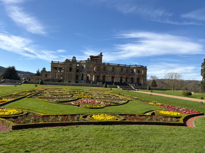The image depicts the ruins of Witley Court, located in the Malvern Hills, United Kingdom, on a sunny day with a blue sky and scattered clouds. In the foreground, a formal parterre garden is meticulously arranged with geometric shapes of manicured hedges, grass, and colorful flower beds. The ruins of Witley Court stand in the background, showcasing remnants of the structure including walls, columns, and a balustrade. The architectural style is classical. The surrounding environment includes grassy slopes and trees. Two people are visible near the far end of the ruins. A gravel path runs to the right of the formal garden.