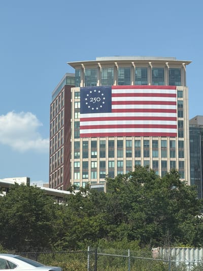 A large American flag is draped across the facade of a modern office building. The flag features a blue canton with white stars and the number "250" in the center. Red and white stripes extend to the right of the canton. The building is made of glass and brick, with multiple floors of windows. Lush green trees are visible in the foreground, partially obscuring the lower portion of the building. A chain-link fence runs across the bottom of the image, with a silver car visible behind it. The sky is a clear blue with a few wispy clouds. The image was taken in Huntington, United States of America.