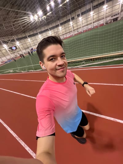 A man with dark hair and a pink t-shirt runs on an indoor track. He's taking a selfie, with his right arm extended towards the camera, wearing a black wristwatch and a white earbud. The track is a reddish-brown with white lane markers. The area next to the track has netting with people in the background. The building has a high ceiling with exposed metal structures and bright lights. He is wearing blue and white gradient shorts with black compression shorts underneath.