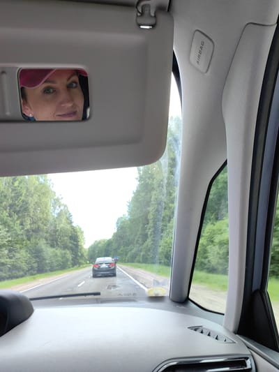 The image captures an interior view from a car, looking forward. In the foreground, the dashboard is visible, along with the windshield. The driver's side sun visor is flipped down, revealing a mirror that reflects the face of a woman wearing a pink baseball cap and earrings. She has blue eyes and is smiling.
Outside the car, the scene is a paved road lined with lush green trees, suggesting a rural or suburban setting during daytime. The weather appears overcast, with a bright sky visible between the trees. Ahead on the road, another car is driving in the same direction.
On the car's interior, above the windshield, a panel is visible with the word "AIRBAG" printed on it, indicating a safety feature of the vehicle. The overall mood is casual and relaxed, possibly during a road trip or everyday commute. The location context is Ilyanskiy selskiy Sovet, Belarus.