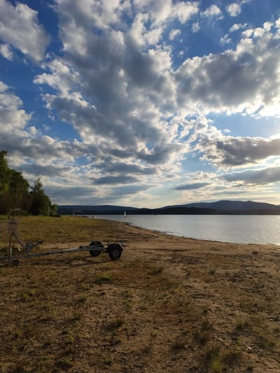 The image depicts a serene lakeside scene at dusk, near Radslav, Czechia. In the foreground, a boat trailer rests on a sandy shore dotted with sparse grass. To the left, a "CAMPING" sign is partially visible, mounted on a pole. The lake stretches out to the horizon, with dark, silhouetted hills in the distance. The sky is a dramatic canvas of blue and white clouds, with the sun casting a warm, golden light from the right, suggesting late afternoon or early evening. Several sailboats are visible on the water, adding a touch of activity to the tranquil setting. The overall impression is one of natural beauty and peaceful recreation.