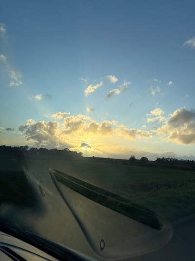 The image, taken from inside a vehicle, shows a view of the sky and landscape near Ruyton-XI-Towns, United Kingdom. The sky is predominantly blue with scattered, fluffy white clouds that are illuminated by the setting sun, which is partially obscured by the clouds near the horizon. Sun rays emanate from behind the clouds. Adjacent to the sky, on the right side of the image, is a field or agricultural area with a line of trees visible along the horizon. The interior of the vehicle, specifically part of the dashboard and possibly the windshield, is visible in the lower right corner, casting a reflection and obstructing some of the view. No people are directly visible. The scene depicts a natural environment at sunset.