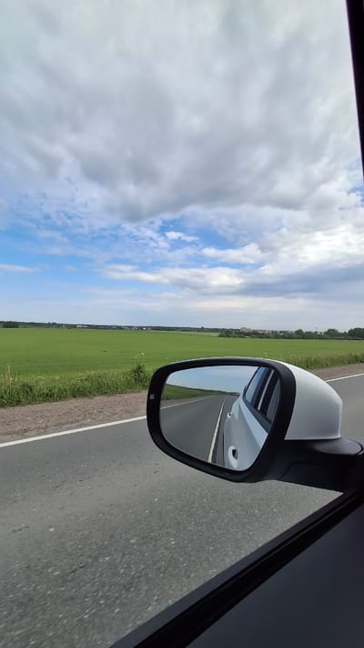 This image captures a wide-angle view from inside a moving vehicle, looking out the passenger side window during daytime in Pigelevo, Russia.
The primary subject is the car's white side mirror, which occupies the mid-right portion of the frame. The mirror's dark trim frames a reflection of the asphalt road receding behind the car, along with a partial view of the car's white body. To the immediate right of the mirror, a dark section of the car's interior or door frame is visible.
Beyond the car, the landscape is dominated by a vast, bright green field stretching horizontally across the middle of the scene, extending towards a distant horizon line where faint trees and structures can be discerned. The field borders a grey asphalt road, which features a clear white line marking its shoulder on the right.
The sky occupies the upper half of the image, presenting a mix of bright blue patches and scattered, fluffy white and light grey clouds, indicating partly cloudy weather. No people or visible text are present in the image. The scene conveys a sense of serene travel through an open, rural landscape.