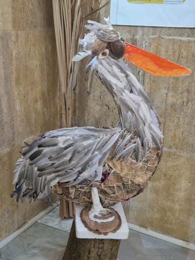 The image shows a hanging sculpture of a bird, likely a waterfowl, located at the Srebarna Lake visitor center in Srebarna, Bulgaria. The sculpture is made of various materials including feathers, shells, an orange paper cutout resembling a beak, and pieces of fabric. The bird's head is constructed from seashells and has an orange paper beak. The neck and body of the bird are draped with feathers, some gray and some white. The body's base appears to be wrapped in a patterned fabric with inscriptions. The sculpture is mounted onto a white foam board to a piece of driftwood. The background consists of a light brown textured wall. Also visible is a straw structure to the side of the bird.