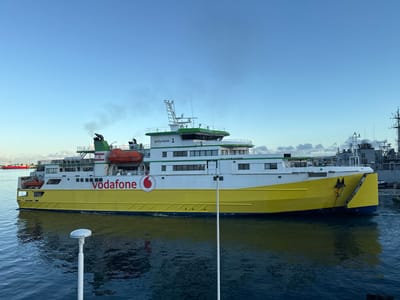The image displays the passenger and vehicle ferry "polynésie 1" in the harbor of Papeete, France. The vessel features a prominent yellow hull and a white superstructure with green accents. The "Vodafone" logo and name are visible in red on the white mid-section of the hull. Black exhaust smoke emanates from two green-topped funnels located towards the stern. Two orange lifeboats are secured on the upper deck. The ferry is positioned on dark blue water, showing reflections of its hull. In the background to the right, a grey naval vessel, possibly a frigate or destroyer, is docked. A red vessel is partially visible in the far-left background. The sky overhead is light blue with scattered white clouds visible in the distance. A white pole-like structure is partially visible in the immediate foreground.