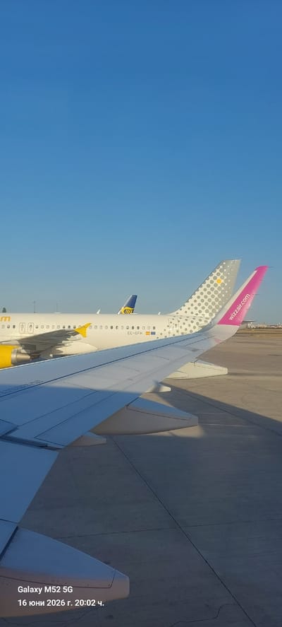 This is a photograph taken from an airplane window, looking out at other aircraft on the tarmac at Manises Airport in Valencia, Spain. The sky is a clear blue, and the sun is casting shadows. In the foreground, the wing of the photographer's aircraft is visible, extending diagonally across the bottom of the frame.
Behind the foreground wing, two other aircraft are parked. The aircraft in the middle has a white fuselage with a yellow wingtip and the registration "EC-OFH" visible. Its tail is partially obscured by the aircraft to its right. The aircraft to the right has a white fuselage with a distinctive grey and white dotted pattern on its tail, and the "wizzair.com" logo is clearly visible in pink.
The airport tarmac is visible in the lower portion of the image, appearing as a concrete surface. The overall impression is one of a sunny day at an airport, with several commercial airplanes lined up. The image also contains metadata in the bottom left corner indicating "Galaxy M52 5G 16 юни 2026 г. 20:02 ч." which translates to "Galaxy M52 5G June 16, 2026, 8:02 PM".
