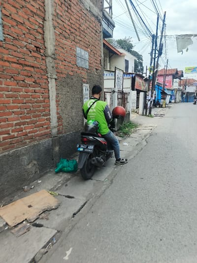 A delivery driver in a green Gojek shirt sits on a black motorcycle on the side of a narrow street. He holds a red helmet. The motorcycle license plate reads "D 3647 ABB." The street is lined with buildings, some brick, and others brightly painted with various signs. Overhead, there are many power lines. A green mesh bag and a piece of cardboard are visible next to the curb. The scene is taken during the day.