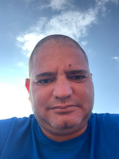 A close-up, low-angle self-portrait of a middle-aged Caucasian man with a shaved head, light stubble, and a mole on his forehead. He is wearing a blue t-shirt. The background shows a bright, blue sky with scattered clouds. The location cannot be determined from this image and there is no indication in the image that it was taken in Zetiovo, Bulgaria.