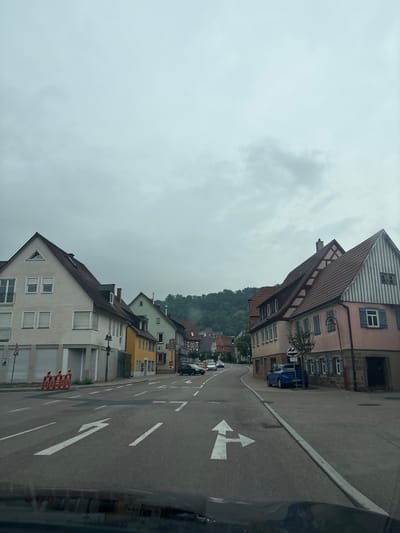 This image captures a street scene in Untermünkheim, Germany, under an overcast sky. The perspective is from inside a vehicle, looking forward and slightly to the right. The road features lane markings, including a white arrow indicating a turn to the left or go straight, and another arrow pointing to the right.
On the left side of the street, there is a light-colored building with multiple windows and closed shutters. Further down the street on the left, there's a yellow building and other structures with reddish-brown roofs, consistent with traditional German architecture. Orange construction barriers are visible on the sidewalk in front of the light-colored building.
On the right side of the street, a building with a pink facade and a red-tiled roof is prominent. This building has several windows, some with blue shutters. A street lamp is positioned in front of it. A blue car is parked on the right side of the road, behind a small tree.
In the background, a wooded hill rises, suggesting the town is situated in a valley or near a natural landscape. Several cars are visible on the road in the distance. The overall impression is a quiet, residential or mixed-use street in a German town.