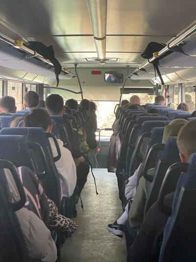 The image displays the interior of a passenger bus or coach, viewed from the rear, looking forward. Rows of dark blue fabric seats with light grey backs are visible, occupied by numerous individuals, mostly seen from behind. A central aisle separates the seating on the left and right. Overhead luggage racks, constructed from metal bars and dark fabric netting, extend along both sides of the ceiling; a black backpack is stored on the left-hand rack. A long, silver-colored grab rail traverses the ceiling. Above the front windshield, a digital display with red numerals is visible, alongside a rearview mirror reflecting a partial image of an individual. Natural light illuminates the interior through side windows, revealing an exterior landscape dominated by trees and foliage, indicating travel through a natural or wooded area. The vehicle floor is light grey. Passengers are seated and oriented forward, wearing various clothing items including hoodies, jackets, and an item with a leopard print. A person's right foot, wearing a white sock and a black and white sneaker, is visible in the right foreground. No specific landmarks are discernible within the image to confirm the exact location within Torbalazhite, Bulgaria.
