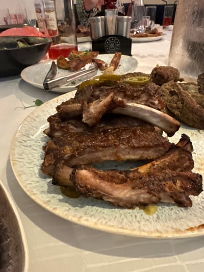The image shows a plate of ribs served on a patterned plate, with other dishes and utensils visible in the background. The ribs appear to be cooked and have a slightly charred appearance. There are also some green vegetables and a dollop of what looks like mustard or a similar condiment on the plate. In the background, there are other food items, glasses, and cutlery. A black bag with a circular logo is also partially visible. The overall setting suggests a meal, possibly in a restaurant or at a gathering. The lighting is warm, suggesting it might be indoors during the day or evening. There is no clear indication of the time of day, weather, or any specific emotions. No visible text is clearly legible.