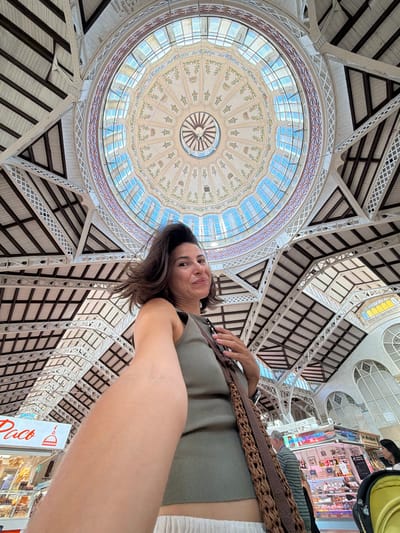 The image shows a woman taking a low-angle selfie inside a building with a large, ornate dome and intricate architectural details. The dome is made of glass panels, allowing natural light to filter in, and is decorated with floral motifs. The ceiling features a complex network of beams and supports, suggesting a grand public space like a market or exhibition hall. The woman is smiling and looking at the camera, with her right arm visible across the frame. She is wearing a green tank top and a woven strap over her shoulder. In the background, stalls or shops are visible, with some signs and displays of goods. The overall impression is one of a bright and lively interior.
A visible sign in red and white text reads "Paco". The context suggests this may be a market or commercial area in Alboraia, Spain. The lighting indicates it is daytime, likely indoors.