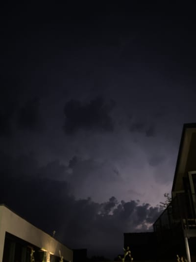 The image depicts a nighttime view of a dark, cloudy sky with hints of lightning flashes. In the foreground, the edge of a modern building with a light-colored facade is visible on the left, and another building structure, possibly a balcony or upper floor with glass railings, is seen on the right. The location is identified as Putten, Netherlands. The sky is predominantly dark with ominous-looking clouds, suggesting an impending storm or ongoing lightning activity. The lighting is dim, consistent with nighttime conditions, and the scene is devoid of any visible people or animals. The overall impression is one of a dramatic weather event occurring over a residential or architectural area.