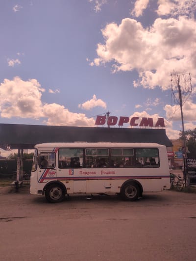 A white bus with red and blue trim is parked on a dirt road in front of a building with the word "ВОРСМА" (Vorsma) in large red letters across its facade. The bus has several windows, and people can be seen inside. The building appears to be a commercial establishment, possibly a bus station or a store. There is a telephone pole with wires on the right side of the image, and a bicycle is leaning against it. Several posters are visible on the wall of the building to the right of the bus. The sky is blue with scattered clouds, and the scene appears to be in an outdoor urban setting.