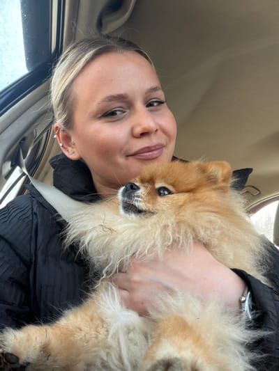 The scene is located inside a vehicle in the city of Makarovo, Russia. A female subject, with light blonde hair pulled back, occupies a passenger seat, facing the camera with a subtle smile. She wears a dark, textured jacket, secured by a light-colored seatbelt that crosses her torso. Her left ear displays multiple piercings: one helix, one tragus, and three lobe piercings. A silver-colored bracelet or watch is visible on her left wrist. She holds a small, fluffy Pomeranian dog, primarily reddish-blonde and cream in color, with both hands. The dog's head is slightly raised and turned towards the viewer's left, its dark eyes looking slightly upwards, and a small pink tongue is visible. The immediate environment is the interior of a vehicle, characterized by a light-colored headliner and a blurred window background indicating daytime outdoor conditions.