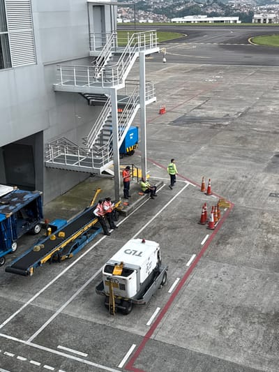 This image depicts an airport ground operations area in the city of AMCO, Area Metropolitana Centro Occidente, Colombia, under overcast conditions.
The scene is dominated by a grey, multi-story building on the left, featuring horizontal louvered windows on its upper section and an attached exterior metal staircase with multiple landings and railings. Below the staircase, a small blue utility cart is partially visible.
The concrete tarmac displays various white and red painted lines and markings. A line of orange traffic cones marks a path or designated area on the right, with a smaller cluster of cones adjacent to it.
Several pieces of ground support equipment are present. On the far left, a blue luggage container is partially visible. Adjacent to it is a blue luggage conveyor belt system, currently stationary, with several yellow items resting on its black belt. To the right, a white ground power unit (GPU) labeled "TLD UPE 207" with a yellow front attachment is parked.
Six individuals, presumably airport ground staff, are visible.
* Two individuals, wearing white shirts and dark pants with reflective vests (pink/orange), are positioned next to the luggage conveyor, one leaning on it, the other standing. Both appear to be holding clipboards or documents and engaging in a discussion.
* Near the staircase base, three individuals are situated by a low concrete barrier. One, in an orange shirt and dark pants with a reflective vest, stands holding a mobile phone. Another, in a green shirt and dark pants with a reflective vest, is seated on the concrete barrier. The third, wearing a yellow/lime green reflective vest over a dark shirt, walks away from the group towards the right side of the frame.
* A surveillance camera is mounted on a pole extending from the staircase structure, overlooking the tarmac.
In the background, a concrete runway or taxiway with green grass verges is visible. Beyond that, distant hills covered with residential buildings extend across the horizon.