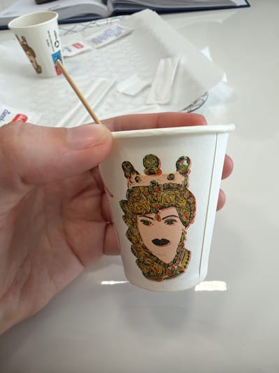 A person's hand holds a small white paper cup with a cartoon drawing of a person in ornate clothing. A wooden toothpick is inserted into the top rim of the cup. A second similar paper cup is on a white surface, which has packaging with the word "Zucchero". A white book is in the background. The location is Scordia, Italy.