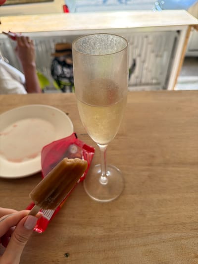 The scene is set at a wooden table within an establishment in บ้านท่าฉัตรไชย, Thailand. In the immediate foreground, a person's left hand, with light pink manicured nails and a tattoo on the thumb, holds a partially consumed, brown-colored popsicle on a wooden stick. The popsicle's red plastic wrapper rests beneath it on the table. To the left of the popsicle, a white, round plate is visible, showing red residue from a previously consumed item. To the right, a champagne flute, approximately one-third full with a pale yellow, effervescent liquid, stands on the wooden surface. The glass has visible condensation and minor surface residue. The background is out of focus, revealing another individual on the left, wearing a light-colored top, with their arm raised towards their mouth, holding a red object. A gold ring is visible on this person's left ring finger. Further right in the background, a blurry figure wearing a straw hat and a dark patterned shirt is discernible behind a counter-like structure with vertical white slats.