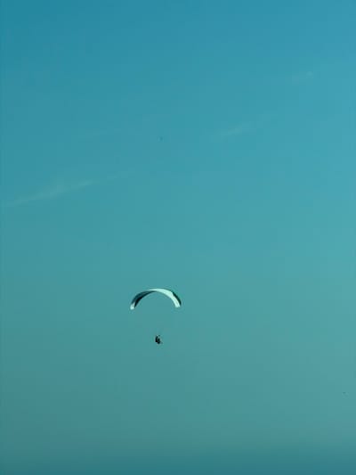 In this media file, a lone paraglider is captured mid-flight against a clear, light blue sky. The paraglider, a person strapped into a harness, is being propelled by a large, crescent-shaped paraglider wing. The wing is predominantly white with dark accents along its edges. The scene is characterized by a vast expanse of sky, suggesting an open-air activity.
The paraglider is positioned in the upper right quadrant of the image, with the wing taking up a significant portion of the right side. The figure of the paraglider is small relative to the wing, highlighting the scale of the equipment. There are a few faint, wispy cloud formations or atmospheric streaks visible in the sky, particularly on the left side. Additionally, two very small, dark specks are present in the sky, likely distant birds or possibly other airborne objects.
The lighting suggests it is daytime, with the sun likely high in the sky, creating a bright and evenly lit scene. There are no distinct shadows or harsh lighting, indicating pleasant weather conditions. The overall impression is one of freedom and exhilaration associated with paragliding. The location context of Ivanovići, Montenegro, implies a setting that could offer scenic views from above, though these are not visible in the frame. No text is visible in the media file.