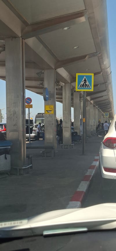 The image depicts an outdoor scene at what appears to be an airport terminal or a similar transportation hub in an unknown city in Israel. The scene is viewed from a vehicle, with the foreground showing the car's windshield and dashboard.
The main area of the image is a covered walkway supported by thick, concrete pillars. These pillars have a weathered, somewhat stained appearance. The ceiling of the walkway is made of light-colored panels with recessed lighting fixtures.
Several signs are visible. A prominent pedestrian crossing sign, a blue square with a yellow border and a black silhouette of a person crossing a zebra stripe, is mounted on a pole. Another sign, a yellow square with a black outline of a smoking cigarette and the word "SMOKING", is attached to one of the pillars. A circular blue sign is also mounted on a pillar. A red and blue circular sign with text in Hebrew is visible on another pillar.
Several vehicles are present, including cars and what looks like a van or small bus. People are seen standing near the vehicles, some with luggage. One person is standing by the open trunk of a car, and others are walking or waiting. The ground is paved, with a curb marked with red and white stripes in the foreground. The overall lighting suggests it is daytime with a clear sky. The environment appears to be an active drop-off or pick-up area.