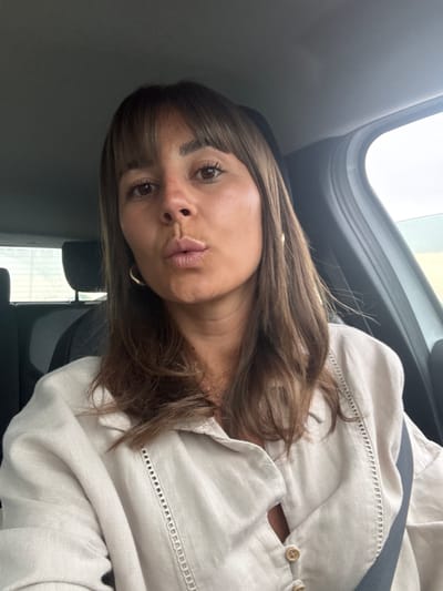 The image shows a close-up, selfie-style shot of a woman inside a car. She has medium-length brown hair with bangs and is wearing a light-colored, button-up shirt. Her lips are pursed, and she is looking towards the camera. Gold hoop earrings are visible. The interior of the car, including headrests and a seatbelt, is partially visible. The background outside the car windows suggests an overcast or bright sky. The context of Les Arcs, France, is not directly depicted in the visual elements of the image.