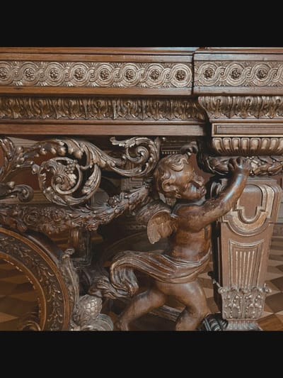 The image shows an ornate wooden carving, possibly part of a large piece of furniture or a decorative architectural element. The carving depicts a cherubic figure with small wings, rendered in dark wood. The cherub is positioned in the lower right of the frame, reaching up with both hands to grasp a decorative urn-like object which is also carved from wood and features foliate details. The cherub is draped in a cloth that wraps around its body. To the left of the cherub, there are elaborate carved acanthus leaves and floral motifs. Above the cherub and the decorative carvings, there is a horizontal band of repetitive circular floral patterns. The overall style suggests a baroque or rococo influence, characterized by intricate detail and dramatic forms. The background is softly blurred, with hints of a checkered floor pattern visible beneath the carving. The specific location within Saint-Aouût, France, is not discernible from the image.