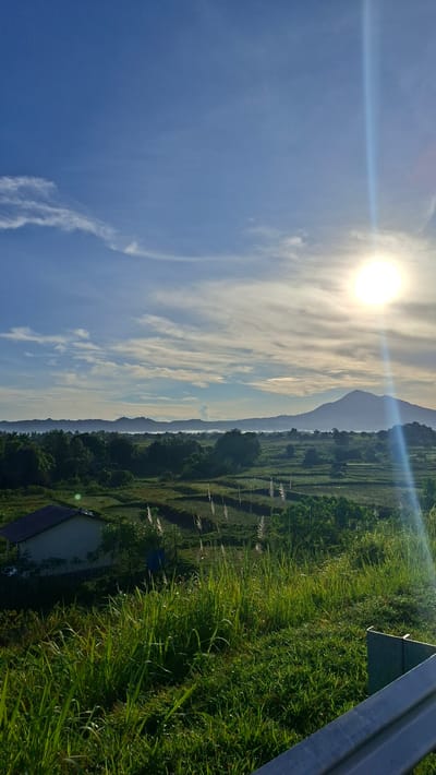 The image depicts a rural landscape bathed in bright sunlight, likely during sunrise or sunset. The foreground features lush green grass and tall reeds, with a metallic railing partially visible on the right side. Beyond the immediate foreground, a small, light-colored building with a dark roof sits nestled amongst trees. The midground is characterized by terraced fields, possibly rice paddies, showing patterns of cultivation and interspersed with vegetation. A dense line of trees forms a natural boundary separating the fields from the distance. In the background, a silhouetted mountain range stretches across the horizon, partially shrouded in a soft haze. The sky is a vibrant blue, dotted with wispy white clouds, and a bright sun with a strong lens flare is visible in the upper right quadrant. The overall impression is one of tranquility and natural beauty in a tropical or subtropical setting, consistent with the environment found in areas like Keureuweungkrueng, Indonesia.