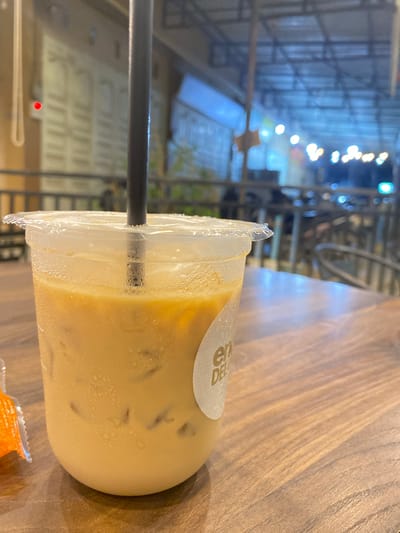 A close-up shot features a clear plastic cup filled with an iced coffee drink, set on a wooden surface. A black straw is inserted into the cup. The cup has a sticker with "EN DELI" visible on it. The background shows an outdoor area with construction scaffolding and streetlights. The blurry background suggests an urban environment. The location is Tanjongkapai, Indonesia. An orange package is partially visible near the bottom edge of the frame.