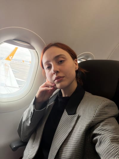 A female individual is depicted inside an airplane cabin, seated in a dark-colored passenger seat next to a window. Through the window, an aircraft wing with a distinctive yellow Pegasus Airlines logo on its tip is visible, indicating the setting is an airport tarmac. This location is identified as Sabiha Gökçen International Airport (SAW), which is geographically adjacent to Tuzla, Turkey. Other aircraft and airport infrastructure are discernible in the distance outside. The individual has reddish-brown hair, pulled back, and wears a black turtleneck under a grey-and-black checkered blazer with solid black lapels. Her right hand, featuring light pink nail polish, is positioned with fingers resting against her jawline. A small stud earring is present in her right ear. She is looking directly at the camera. The airplane appears to be stationary on the ground.