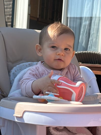 A baby is sitting in a high chair with a drink bottle. The high chair is white and beige. The drink bottle is red and white with a white handle. The baby is wearing a pink long-sleeved shirt. The background shows a window and outdoor scenery. There is no information provided about the city of Veghel, Netherlands.