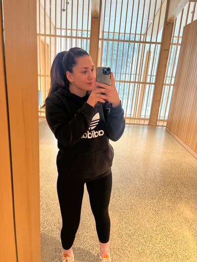 An adult female is depicted taking a mirror selfie inside a modern building in Erstfeld, Switzerland, specifically identifiable by its architectural style as likely the Gotthard Base Tunnel Visitor Center. The subject has dark hair pulled into a high ponytail and wears a black Adidas hooded sweatshirt with a white logo, black leggings, light pink ankle socks, and white athletic shoes with orange laces. She holds a light blue/grey cased smartphone in her right hand, directing it towards the mirror to capture her reflection. Her gaze is fixed on the phone's screen. The immediate environment includes a light-colored, speckled terrazzo floor. The background consists of large architectural windows framed by numerous vertical and horizontal natural wood beams, creating a grid-like pattern. Beyond these windows, blurred green foliage and hints of an exterior environment are visible. A plain wooden panel serves as the reflective surface on the left, and a vertically planked wooden wall is present on the right.