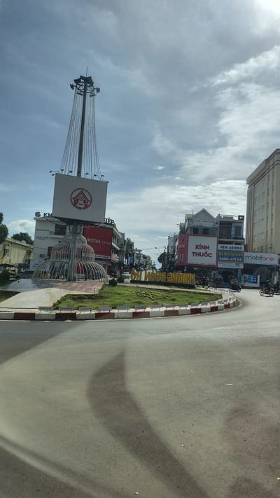 The image captures a street scene in Buon Ho, Vietnam, featuring a roundabout in the foreground. Dominating the scene is a tall, slender tower topped with a large square billboard displaying a red and white logo and text. The billboard is suspended by cables. Below the billboard is an ornamental, tiered metal structure resembling a fountain or abstract sculpture.

The roundabout has a red and white striped curb. Beyond the roundabout, buildings line the street. One prominent building has a "Viettel store" sign. Another building displays "Kính Thuốc" (pharmacy). Further down the street, a large, light-colored building with multiple windows is visible. A sign reading "mobifone" is also present.

In the center of the roundabout, a grass area displays yellow lettering that reads "THÀNH PHỐ BUÔN HÔ" (City of Buon Ho). Several motorcycles are visible on the road, with two riders on one motorcycle and another single rider on a separate motorcycle. The sky is partly cloudy. Shadows of structures, likely from the surrounding buildings and the central sculpture, are cast on the asphalt road.