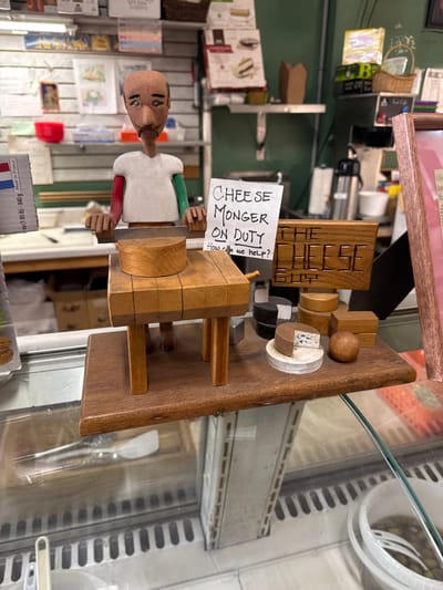A wooden carving of a cheese monger with a sign that says "CHEESE MONGER ON DUTY. How can we help?" sits on a wooden surface next to various wooden cheese-shaped pieces. The scene is in a store, possibly behind a glass display, with shelves of products in the background. A price tag of "ea (1.85 lb)-840 g Made in France" is visible, providing additional context.