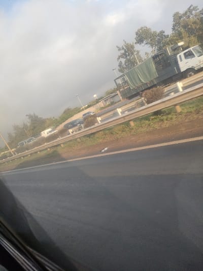The image displays a tilted view of a multi-lane roadway in the city of Theta ward, Kenya, observed from within a vehicle. The immediate foreground shows a dark asphalt road surface with a faint light-colored line delineating a slightly lighter shoulder, where a small white object rests. A silver-grey guardrail runs across the midground, separating the main roadway from a parallel lane or service road. Along this parallel section, a queue of vehicles is visible, including a prominent white truck with a dark chassis and a green canvas covering its cargo area, positioned on the right. Additional passenger vehicles, including a white van and darker cars, are present further along this lane. The background features scattered green trees, multiple tall streetlights, a low-lying light-colored building, and a white water tower with a conical roof. The sky is uniformly overcast with light grey clouds. No individuals are discernible within the scene.