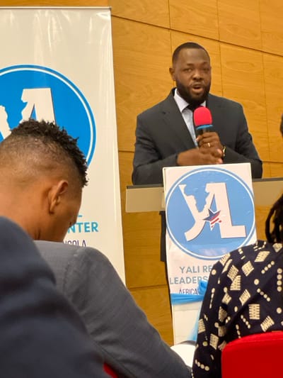 An indoor scene captures a presentation or discussion at a YALI (Young African Leaders Initiative) event in Luanda, Angola. The main subject is a man of African descent, dressed in a dark suit and white shirt, speaking animatedly into a red and black microphone. He has a short beard and mustache, and a ring is visible on his left hand.
Next to the speaker, a light blue and white circular sign displays the YALI logo, which includes a map of Africa and a star. Text on a banner beneath this sign reads "YALI REGIONAL LEADERS CENTER AFRICA," though parts are obscured. Another person, with dark, patterned attire and a red shoulder strap, is partially visible seated to the speaker's right.
In the foreground, the back of another individual's head, featuring short, curly hair, suggests an audience member. A large banner, similar to the one beside the speaker, is also visible in the lower part of the frame, with partially legible text including "CENTER" and "ANGOLA," further indicating the event's location and nature. The background consists of light wooden wall paneling. The scene conveys an atmosphere of engagement and leadership development.