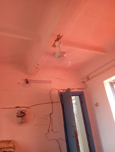 The image depicts an interior scene, likely a room in Vostochnoe, Kyrgyzstan. The room has white walls and a white ceiling with visible signs of wear and tear, including cracks and some damage to a wooden beam supporting a ceiling fan. A white ceiling fan with three blades is mounted on the ceiling. A fluorescent light fixture is attached to the wall, with a light bulb visible. Electrical wires are routed across the ceiling and walls. A blue wooden door is ajar, revealing a glimpse of another room beyond. On the wall to the left, there is an electrical meter. A white electrical panel is mounted below it. In the lower left corner, a stack of folded fabric, possibly blankets or bedding, is visible. A window with a light colored frame is visible on the right side of the image. The lighting in the room is warm, suggesting it might be illuminated by artificial light or influenced by external light conditions.
