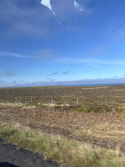 The image depicts a flat, open landscape under a bright blue sky with scattered clouds. In the foreground, dry, brown grass and sparse vegetation cover the ground, with a dark, indistinct area on the lower left possibly indicating a road. A wire fence with thin, vertical posts runs horizontally across the mid-ground. Beyond the fence, the terrain shifts to a mix of low-lying green and reddish-brown vegetation, appearing somewhat mossy or heath-like. In the distance, a body of water, likely the ocean, stretches to the horizon. Further back, a range of low, undulating mountains is visible, some with patches of snow or light-colored vegetation. The overall impression is one of a vast, sparsely populated rural area, characteristic of the landscape around Blönduós, Iceland. There are no people or specific identifiable structures visible in the image. The scene appears to be captured from a moving vehicle due to the slight motion blur in the foreground grass.