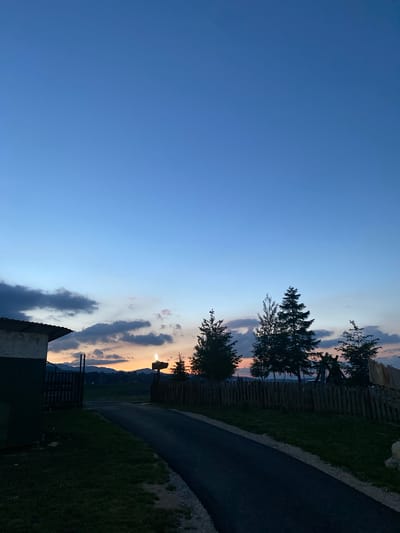 The image captures a dusk scene in the city of Javorje, Montenegro. A clear blue sky transitions to a softer hue near the horizon, where clouds are tinged with the warm colors of sunset. Silhouetted against this backdrop are several coniferous trees of varying heights, and in the foreground, a dark asphalt road leads away from a small building on the left. The building has a dark green section on its side and a partial view of a fence. A rustic wooden fence runs along the right side of the road, and further back, the faint outline of mountains can be seen. A light fixture on a pole stands near the center, emitting a faint glow. The overall atmosphere is peaceful and serene, typical of an evening in a rural or semi-rural setting.