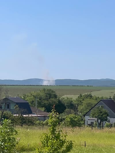 In a rural setting, likely within or near the city of Zarya, Russia, a panoramic view captures a distant plume of smoke rising from behind a range of green hills under a clear, pale blue sky. In the foreground, a lush field of tall green grass and scattered trees creates a natural border. To the left, a small house with a dark roof and light-colored walls is visible, partially obscured by trees. To the right, another house, also with a dark roof and white walls, sits amongst dense greenery. The landscape suggests a tranquil, possibly agricultural or semi-rural community.