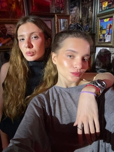 The image shows two young women in a close-up, horizontal shot. The woman in the foreground, positioned at the bottom of the frame, has long, wavy brown hair that falls across her face and chest. She is looking upwards with her mouth slightly parted, her skin appearing flushed and shiny. She is wearing a black garment underneath a grey shirt.
The woman behind her, positioned in the upper half of the frame, has lighter brown hair pulled back and is also looking upwards. Her skin is also shiny, and she has makeup on her eyes and lips. She is wearing a grey t-shirt and has a watch with a metal band and red and yellow accents on her left wrist. A thin purple bracelet is also visible on the same wrist. Her right hand, with a ring on the index finger, is resting on her chest.
Both women appear to be indoors, with a background composed of numerous framed pictures. The frames are of various sizes and colors, with some of the images within them visible. These images depict what appear to be colorful landscapes and possibly cityscapes. The lighting in the scene is warm and appears to be artificial, possibly from overhead lights, casting shadows on the subjects and the background. The overall impression is one of a casual, intimate moment captured between two friends. The setting seems to be a room decorated with many framed artworks. There is no visible text in the image.