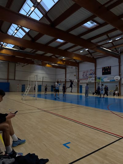 An indoor sports hall in Sennecey-le-Grand, France, is depicted during what appears to be a volleyball game or practice. The facility features a light-colored wooden floor with blue-painted areas marking a volleyball court, delineated by white, black, and red lines. A white referee stand with a ladder is positioned beside a standard volleyball net.
Approximately 15-20 individuals are present. In the immediate foreground, a young male is seated, viewing a smartphone. He wears dark shorts, white socks, and light blue sneakers. On the volleyball court, several male players are actively engaged. One player, wearing a light blue jersey with the number "7", is captured mid-air near the net, appearing to set or hit a volleyball. Another player in a dark jersey is similarly airborne on the opposite side of the net, likely attempting a block or spike. A volleyball is visible in the air above the net.
In the background, two basketball backboards and hoops are mounted on the walls. A digital scoreboard displays "LOC 2" and "VIS 1", with "L" and "1" for sets, indicating a score of 2-1 (local leading 0 sets to 1 set). Banners and posters are affixed to the far wall, one prominently featuring "Sennecey-le-Grand". A group of approximately 5-7 individuals, including adults in casual attire, are standing or observing the activity in the far right background. The ceiling comprises a network of wooden beams supporting a corrugated roof and multiple rectangular skylights, augmented by artificial fluorescent lighting fixtures.