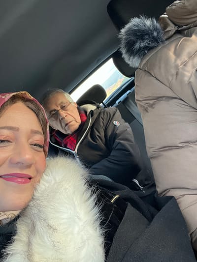 The image, originally oriented sideways, captures a selfie taken inside a vehicle, likely during the daytime. In the foreground, a woman with light makeup and pink lipstick is smiling and looking towards the camera. She is wearing a coat with a prominent white faux fur trim, and part of a patterned headscarf (pink/red tones) is visible.

Behind her, to the left, a man is seated, also looking towards the camera. He wears glasses, a black puffer jacket over a red and black plaid shirt, and has a grey/white furry collar. A round badge or pin with red, white, and blue colors is visible on his jacket, but any text or logo is unreadable.

The setting is the interior of a car or van, with dark upholstery and headrests visible. Through the vehicle's window in the background, a blurred outdoor scene suggests a road or distant landscape. A fluffy black and grey object, possibly a microphone wind muff, hangs from the upper interior of the vehicle on the left. The attire of both individuals suggests cool or cold weather. The woman's smile conveys a positive mood. The specific location of Majmaa Tolba, Morocco, is provided, but no visual cues in the image directly confirm this.