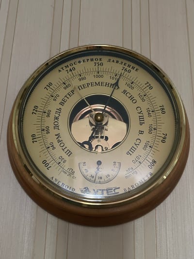 This is a close-up, eye-level shot of an antique aneroid barometer mounted on a lightly textured, pale wall. The barometer is circular with a wooden frame and a brass bezel. The face of the barometer is a cream-colored dial with black markings and a glass cover. The dial displays various atmospheric readings and weather indications in Russian.
The main dial shows pressure readings from 700 to 1070, with smaller increments marked. There are also words indicating weather conditions. On the left side of the dial, in vertical text, are the words "АТМОСФЕРНОЕ ДАВЛЕНИЕ," meaning "Atmospheric Pressure." Along the top arc, the text reads "ЯСНО СУШЬ В. СУШЬ," which translates to "Clear Dry Very Dry." Below this, larger text reads "ВЕТЕР ПЕРЕМЕННО," meaning "Wind Variable." On the lower right arc, there is the word "ШТОРМ ДОЖДЬ," which means "Storm Rain."
There are two smaller dials within the main dial. The larger one on the right has markings from -10 to 50, with smaller tick marks. The smaller dial on the left shows a pointer. The barometer has two hands; a longer, thinner hand pointing to approximately 740 and a shorter, thicker hand pointing to approximately 1010.
The wall behind the barometer has a subtle horizontal stripe pattern. The lighting is bright and even, suggesting it might be daytime. There are no people or other objects visible in the frame, making the barometer the sole focus. The overall impression is of an old, functional instrument, possibly a decorative piece in a home or a historical setting. The text "BAROMETER" and "ANEROID" are also visible on the right side of the dial. The brand "TEC" is also visible on the right side of the dial. The context of Edem, Russia is noted but not visually discernible from the image itself.