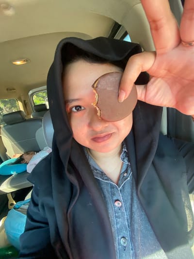 In the interior of a vehicle, a person wearing a black hijab and a denim shirt is holding a broken chocolate cookie in front of their face, obscuring one eye. The person is looking directly at the camera. In the background, a child is asleep on a blue pillow on the back seat of the car. The overall setting appears to be a sunny day, with natural light filtering into the vehicle. The image was captured in Jotangan, Indonesia.